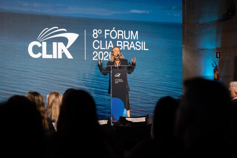 Palestrante em um palco durante o 8º Fórum CLIA Brasil 2023, com tela ao fundo exibindo o logo CLIA e a programação, em evento para público sentado.