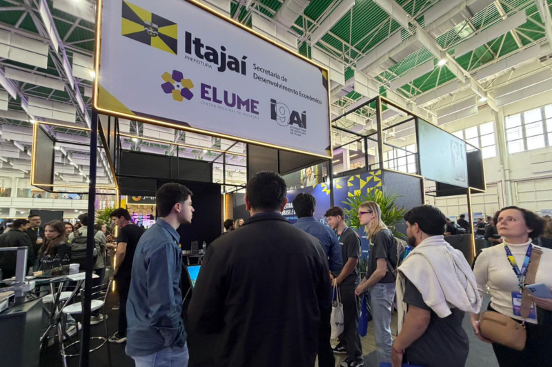 Participantes circulam em um evento em um pavilhão com estandes e telões; ao fundo, banner do Itajaí Secretaria de Desenvolvimento Econômico com as marcas ELUME e GAI iluminadas