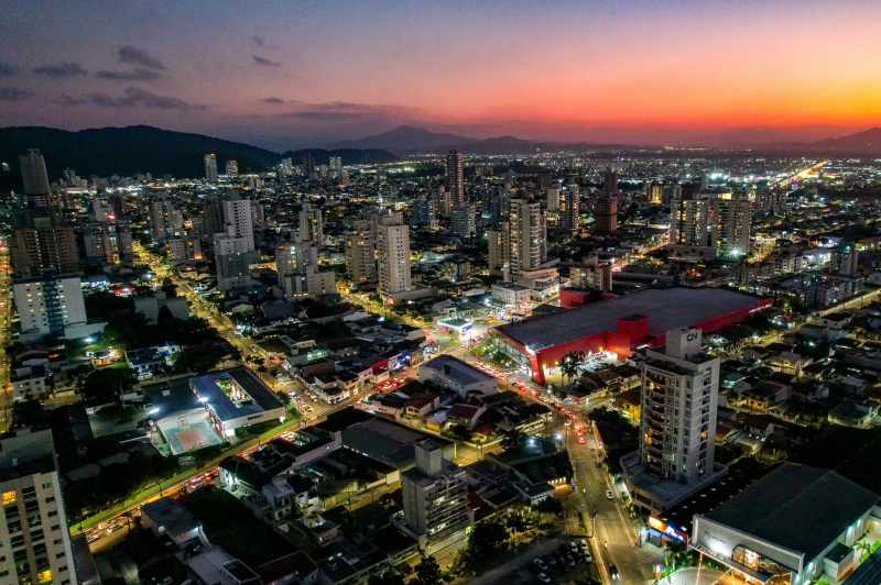 Vista aérea de uma cidade ao entardecer, com edifícios altos, ruas movimentadas e luzes acesas ao fundo, sob céu colorido com tons de rosa e roxo.