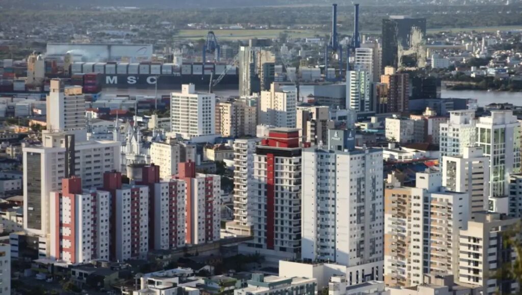 Vista aérea de uma cidade com prédios residenciais e arranha-céus ao fundo, mostrando o skyline urbano e a movimentação da área industrial.