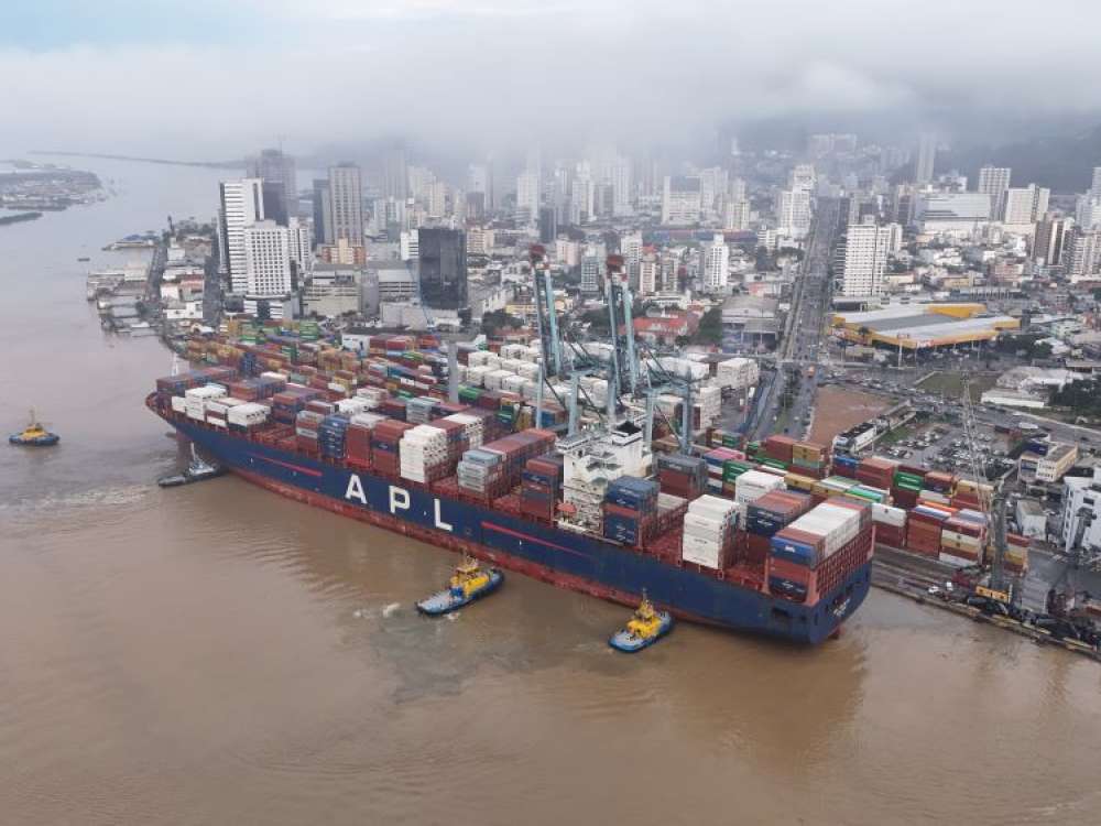 Navio de carga com contêineres no rio em meio a um porto, com o nome “APL” visível e vista da cidade ao fundo, em clima nublado.