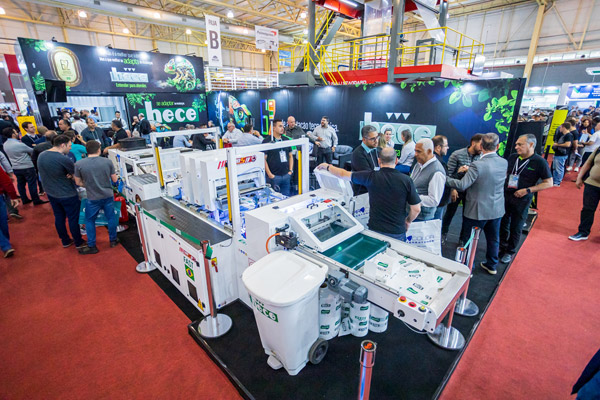 Pessoas visitam estande de feira em ambiente industrial, com equipamentos e maquinário montados sobre bancadas brancas em uma área de exposição com telões ao fundo.