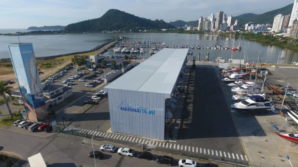 Vista aérea da marina com barcos e iates atracados, prédio de apoio com a placa “MARINATAJAJ” e veículos no pátio, em frente à Baía de um litoral com montanhas ao fundo.