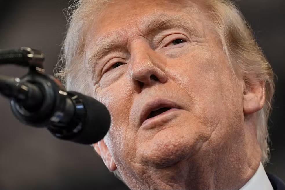 Donald Trump em close, com expressão séria, falando em frente a um microfone com câmera ao lado durante entrevista ou pronunciamento.
