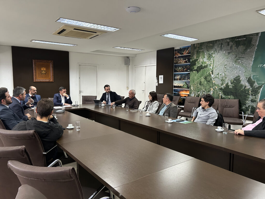 Reunião em sala de conferências com participantes sentados ao redor de uma mesa, falando e analisando informações, com mapas e imagens ao fundo e iluminação de escritório no teto.