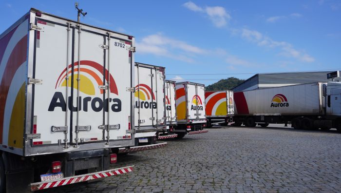 Caminhões de carga refrigerada com a marca Aurora estacionados em pátio, com caçambas traseiras e rodas alinhadas no asfalto e céu azul ao fundo.