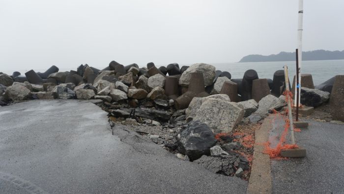 Piso de concreto com pedras grandes e pequenas ao lado do mar, com redes e postes fixados na praia, ao fundo uma enseada e montanhas