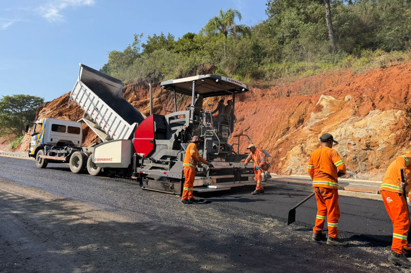 Equipe de trabalhadores de obras realiza aplicação de asfalto com uma máquina niveladora em uma estrada, enquanto caminhão basculante faz o transporte de material em uma área de terraplanagem.