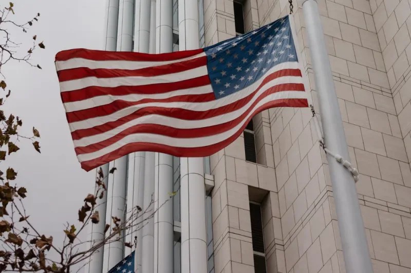 Bandeira dos Estados Unidos tremulando ao vento em frente a um prédio de pedra e colunas, com outra bandeira menor ao fundo, em ambiente urbano.