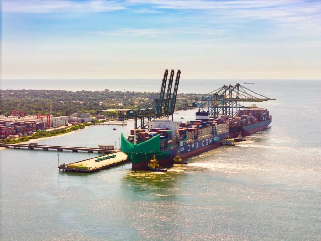 Vista aérea de um navio cargueiro no porto, com contêineres empilhados e o casco verde e azul manobrando no canal ao lado de instalações portuárias e horizonte do mar ao pôr do sol.