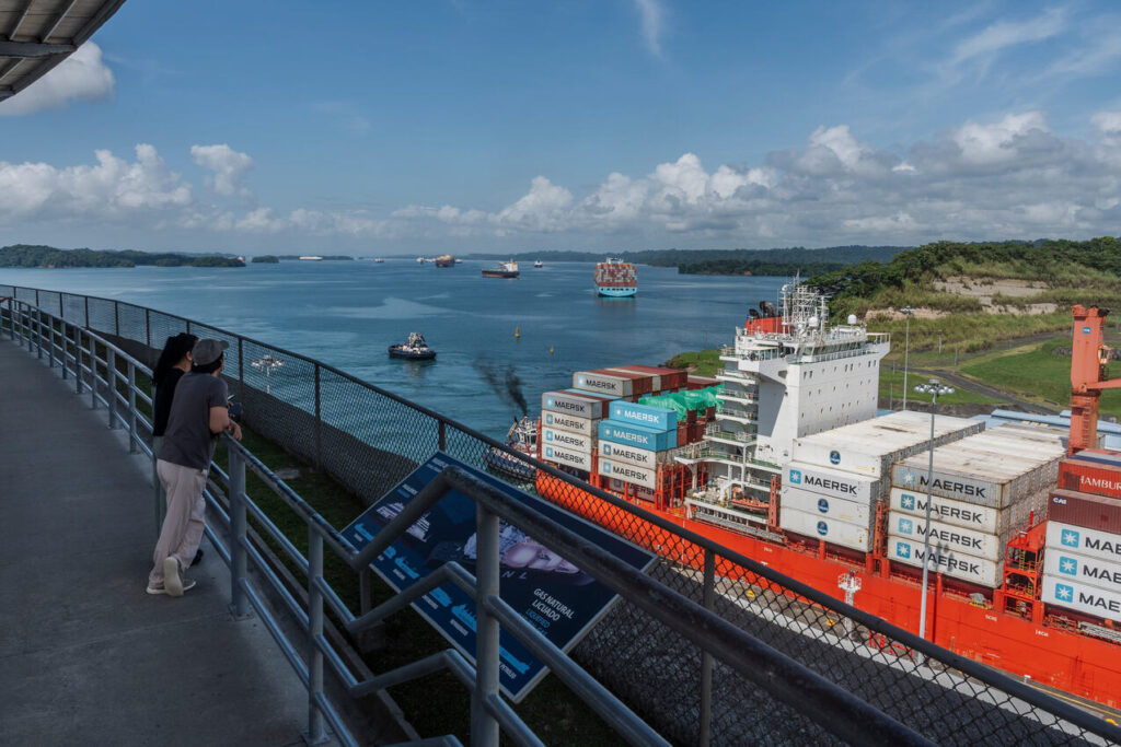 Vista de um terminal portuário à beira de um canal, com navios cargueiros passando e um grande navio com contêineres MAERSK em destaque, além de pessoas caminhando em um passadiço