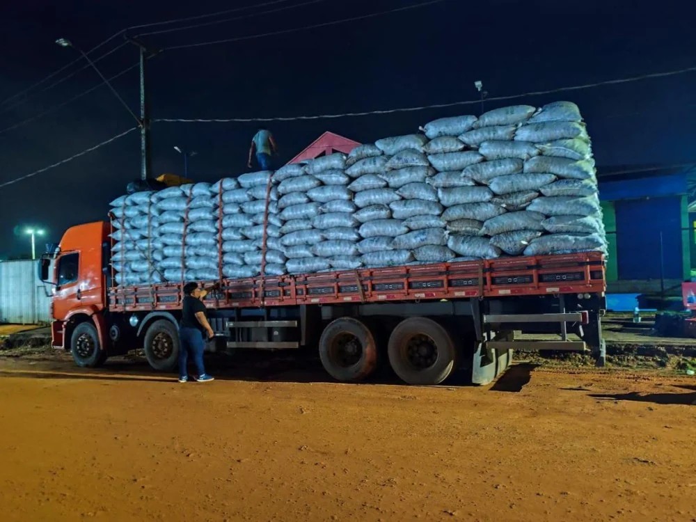 Caminhão carregado com sacos de grãos, empilhados em grande quantidade sobre a carroceria, durante a noite em pátio iluminado, com trabalhador ao lado