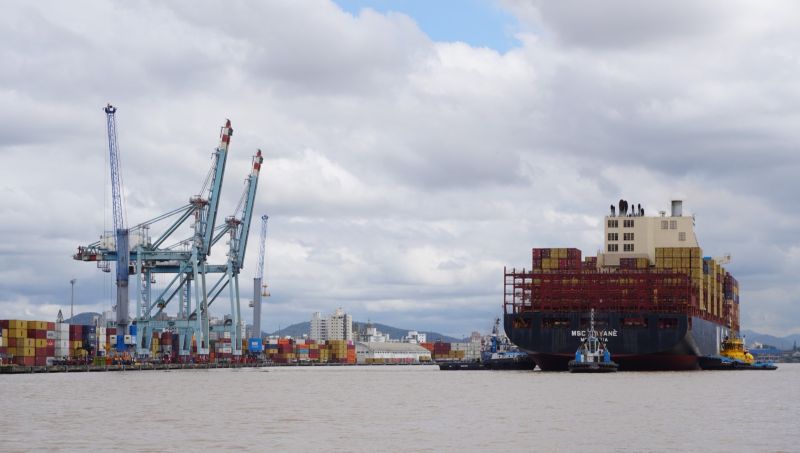 Porto com guindastes e contêineres em um pátio logístico, ao lado de um navio cargueiro grande atracado, com céu nublado ao fundo