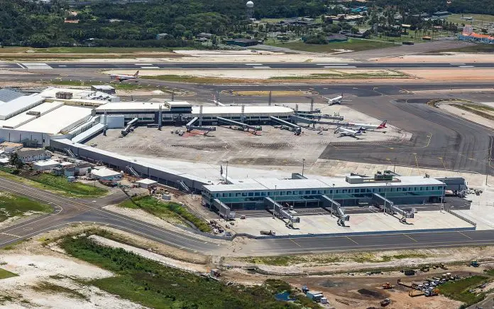 Vista aérea de um aeroporto internacional com pista de pouso e decolagem, pátio de aeronaves, terminal de passageiros e áreas de serviços ao redor, cercado por vegetação e estradas.