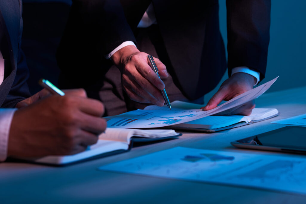 Pessoas em reunião assinando e revisando documentos em uma mesa, com caneta e papéis abertos, em ambiente corporativo com iluminação azul e foco nas mãos.