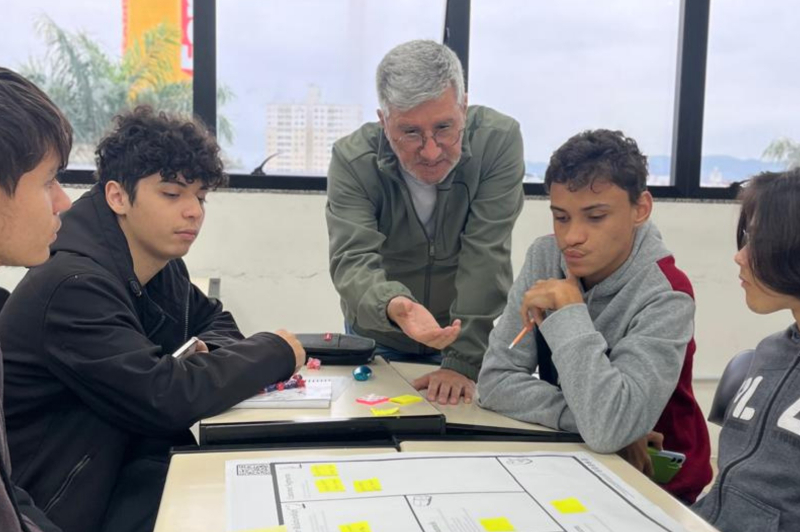 Professor idoso e alunos em sala de aula, trabalhando em uma atividade educativa com mesa e materiais impressos, enquanto ele orienta as crianças.