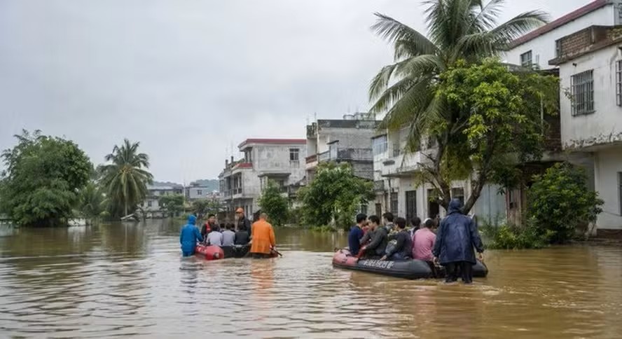 Pessoas em uma balsa improvisada evacuando em área alagada ao lado de casas com palmeiras e céu nublado, durante enchente.