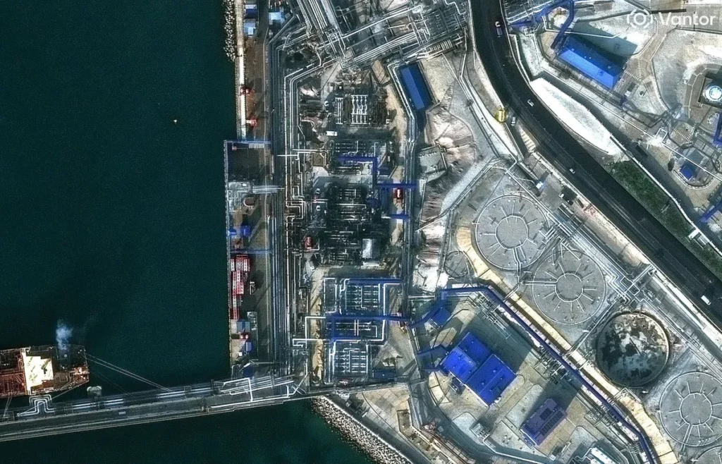 Vista aérea de uma planta industrial petroquímica ao lado do mar, com tubulações, tanques e estruturas metálicas em múltiplas áreas, incluindo pátio de armazenamento e cais