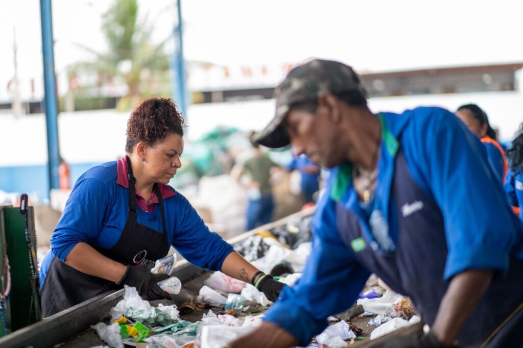 Equipe de coleta seletiva separando materiais recicláveis em uma esteira, com trabalhadores usando luvas e aventais durante o processo de triagem no centro de reciclagem.