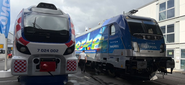 Vista traseira de dois trens ferroviários, um com pintura azul e outro prateado, em área externa ao lado de prédio industrial, com maquinista ao fundo.