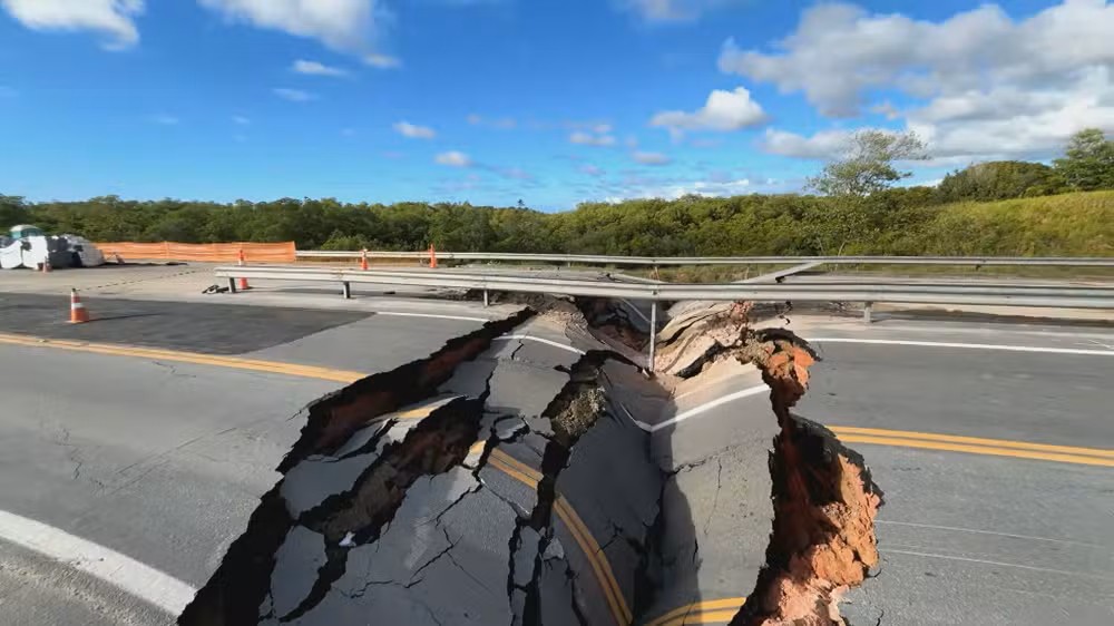 Rachadura e buraco em pista de asfalto em rodovia, com pavimento danificado e sinalização de obra ao fundo, durante inspeção de manutenção