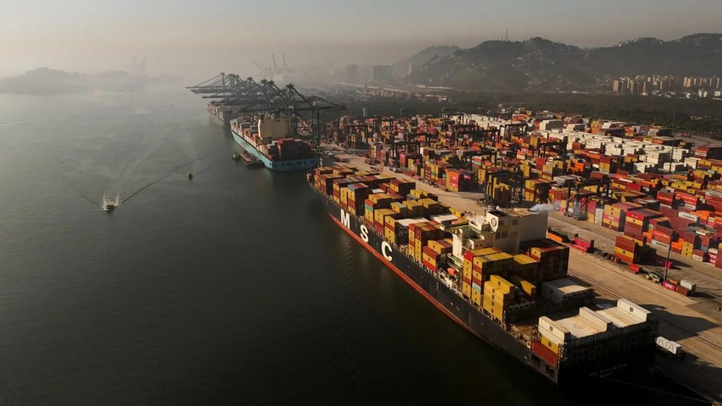 Vista aérea de um porto com grande navio cargueiro ao lado de contêineres coloridos, trilhos de movimentação e paisagem costeira ao fundo