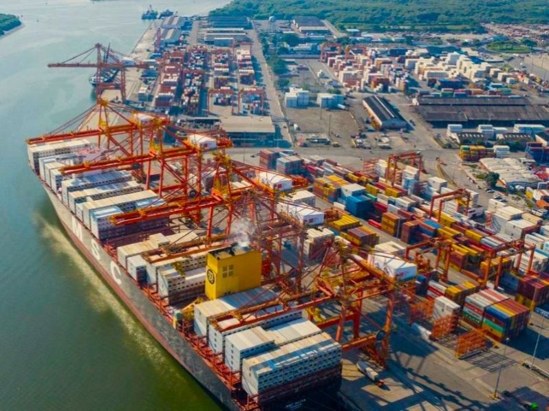 Vista aérea de um grande navio cargueiro no porto com contêineres empilhados e operação de guindastes na área de embarque e desembarque.