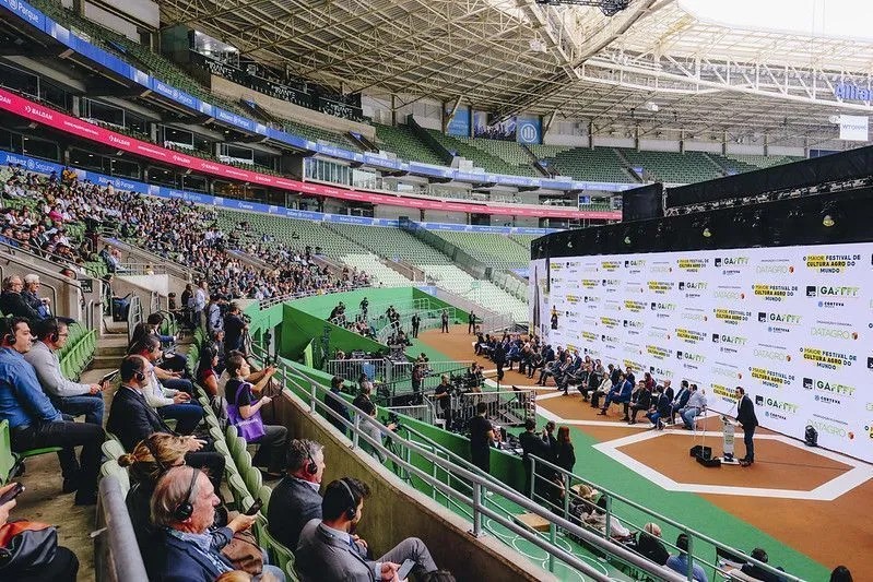 Vista interna de uma arena esportiva durante evento de tênis, com arquibancadas cheias, quadra central e equipe no entorno do torneio.