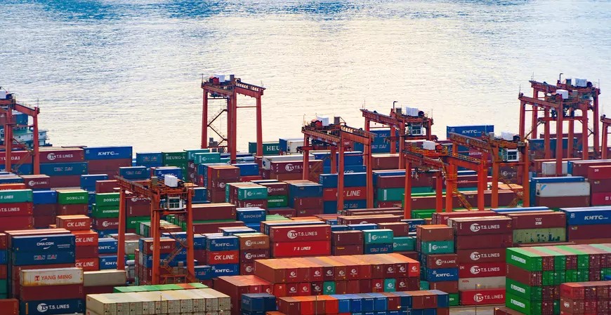 Vista aérea de um grande porto de contêineres com pilhas de cargas coloridas e pórticos industriais ao fundo, com navios e água no horizonte.