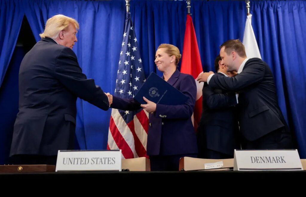Donald Trump entrega um documento a uma mulher em um ato oficial, enquanto um homem segura um envelope ao lado; ao fundo aparecem bandeiras dos Estados Unidos e da Dinamarca e placas “UNITED STATES” e “DENMARK”.