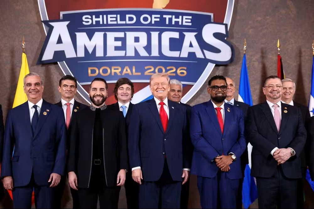 Foto coletiva em frente ao letreiro “SHIELD OF THE AMERICAS DORAL 2026”, com líderes e políticos em trajes formais, em um cenário de encontro internacional.