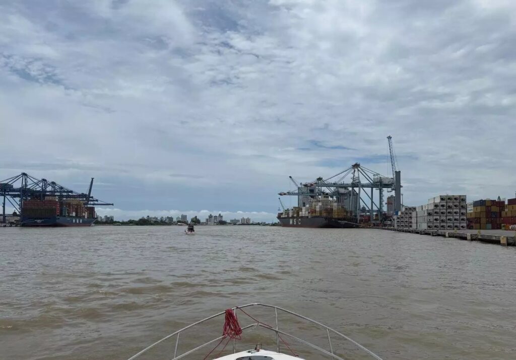 Vista do rio com navios cargueiros e estruturas portuárias ao fundo, com contêineres empilhados e céu nublado, fotografado a partir do convés de um barco.