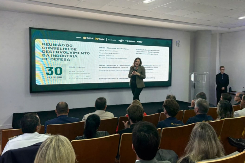 Palestrante em pé apresentando em auditório lotado, enquanto um telão exibe o tema “Reunião do Conselho de Desenvolvimento da Indústria de Defesa” e data de 30 de setembro, em evento corporativo