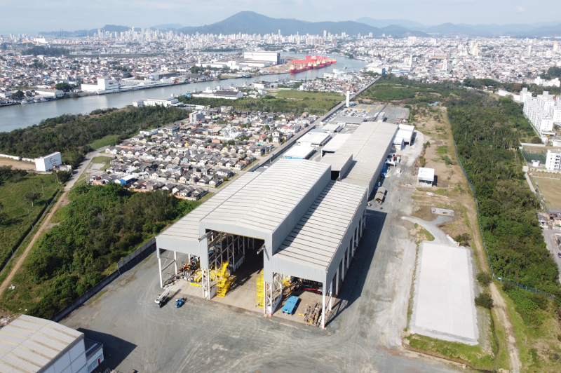 Foto aérea de uma planta industrial com pavilhões metálicos e tanques, cercada por vegetação e bairros residenciais, com vista de um porto e cidade ao fundo.