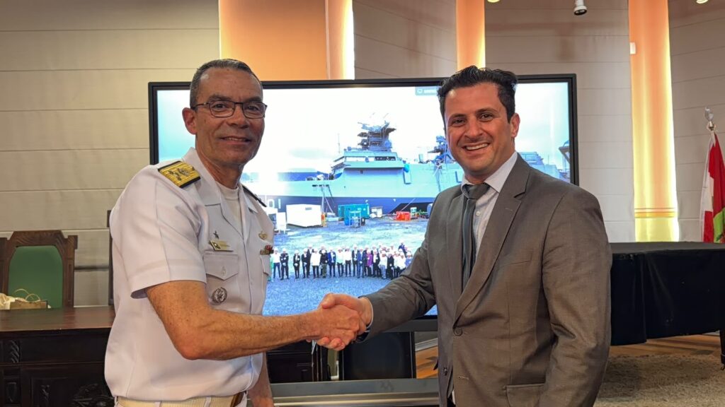 Dois homens, um militar fardado e outro em traje social, fazem um aperto de mãos em ambiente de reunião com tela ao fundo mostrando navio no mar, bandeira e iluminação interna