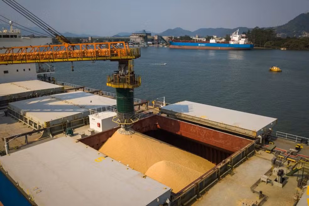 Vista aérea de um navio cargueiro em um porto, com guindaste amarelo e estrutura de carregamento operando em uma baía cercada por montanhas ao fundo.