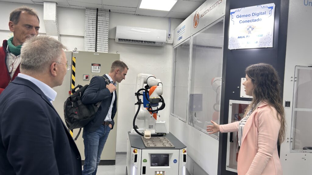 Visita em laboratório técnico com especialistas ao lado de um equipamento automatizado, incluindo um braço robótico e telas de controle, enquanto uma mulher observa o funcionamento na área de testes.