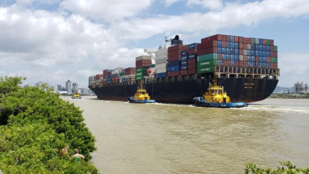 Navio porta-contêineres atracado à margem de um rio, com grandes blocos de contêineres coloridos empilhados no convés e dois rebocadores auxiliando no carregamento. Ao fundo, o céu nublado e a cidade à distância.