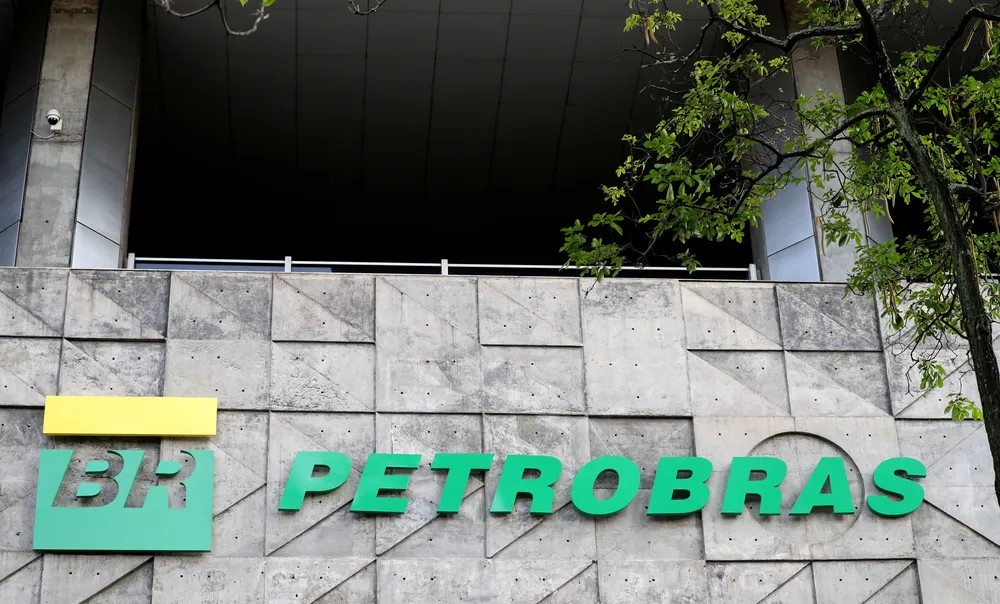 Placa de fachada com o logo “BR” e o texto “PETROBRAS” em destaque sobre parede de concreto, com galhos e folhas verdes ao lado.