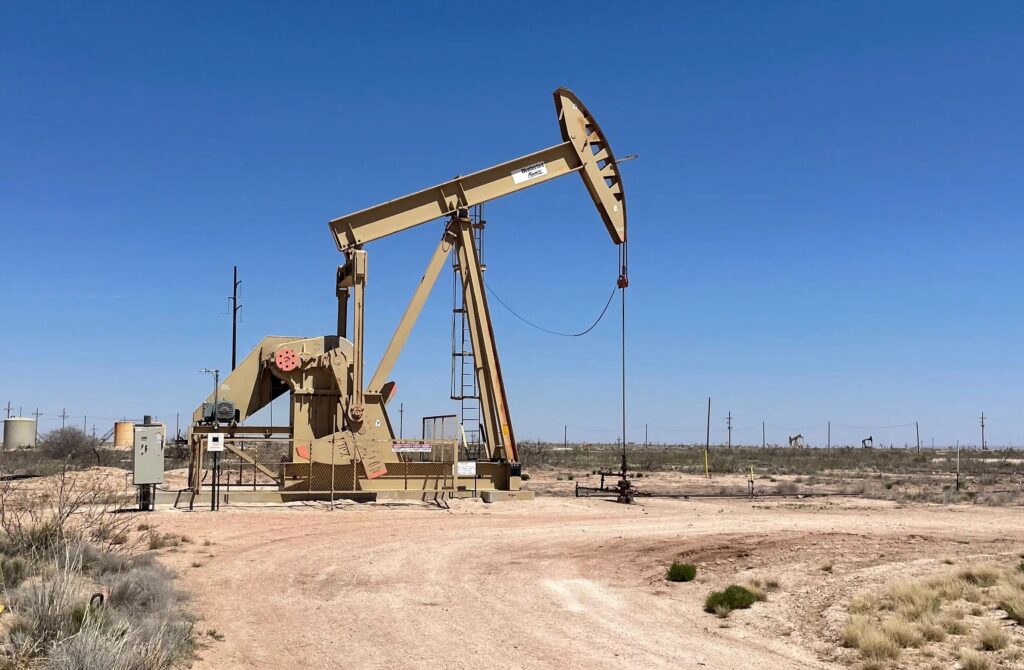 Bomba de extração de petróleo com haste e contrapeso em campo aberto, instalada em um poço no deserto sob céu azul, ao lado de estruturas técnicas.