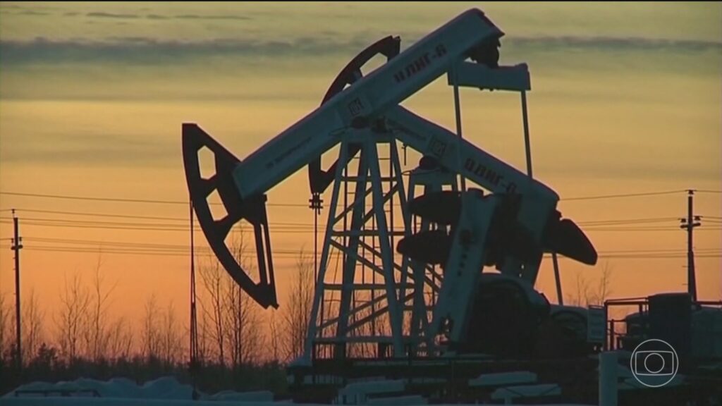 Foto de um equipamento de bombeio de petróleo (bomba de extração) ao pôr do sol, com silhuetas de árvores ao fundo e céu alaranjado, indicando exploração de óleo e gás.