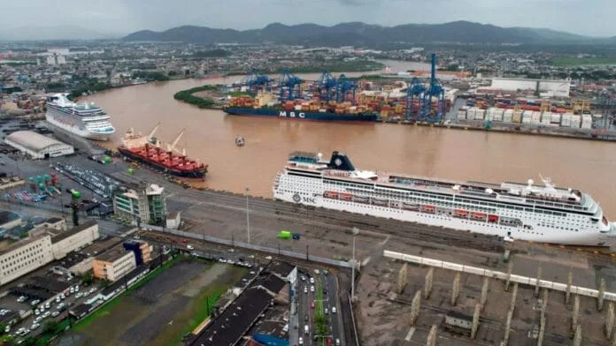 Vista aérea de navios de cruzeiro e carga no porto, com grandes embarcações atracadas ao longo do cais em um cenário urbano e montanhas ao fundo.