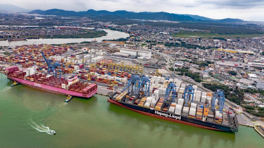 Vista aérea de um porto com contêineres em navios cargueiros e guindastes no cais, com rio ao lado e cidade ao fundo, em cenário industrial logístico.