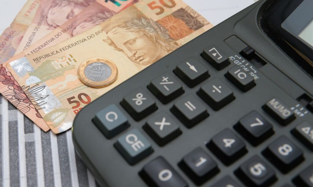 Calculadora financeira com notas de dinheiro brasileiro e moeda de 1 real ao fundo, em cenário de finanças e orçamento.
