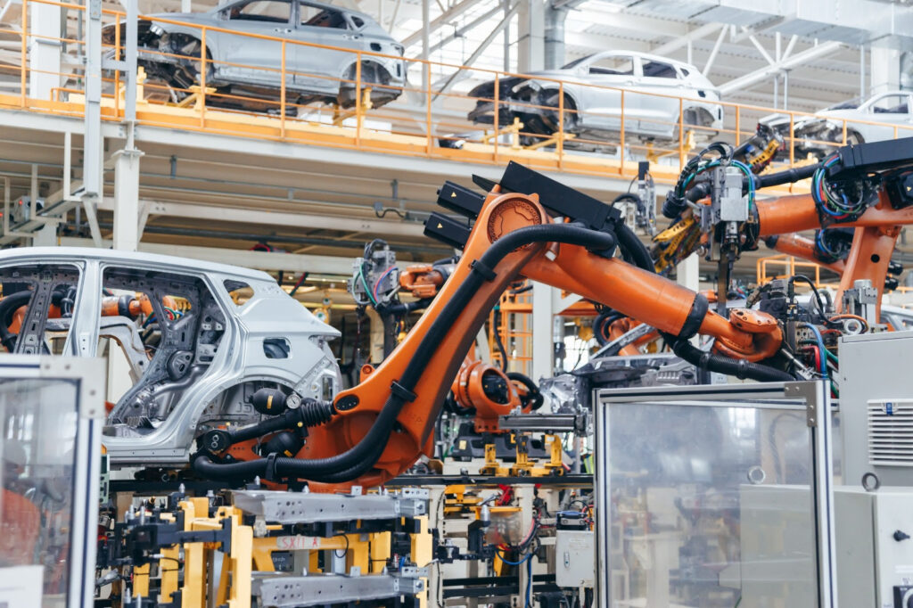Interior de uma linha de montagem industrial com braço robótico laranja, cabos e sensores, além de módulos do carro em uma fábrica moderna