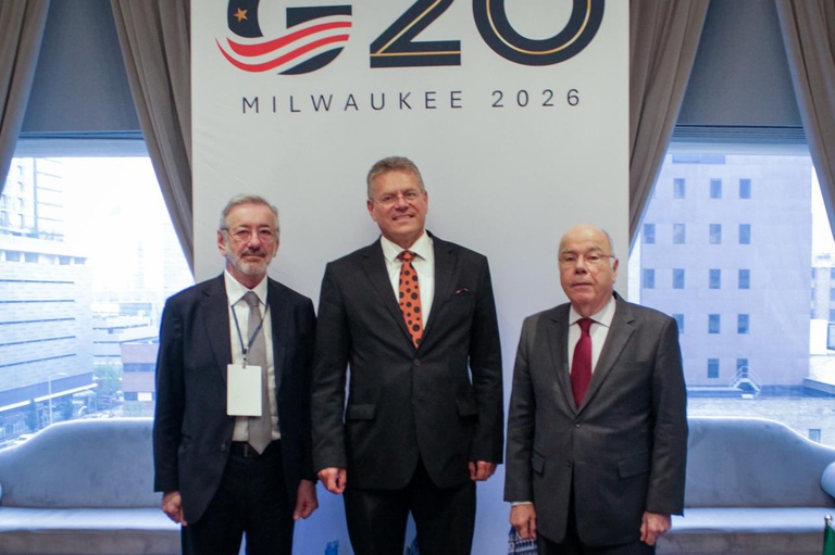 Três homens de terno e gravata posam ao lado de um banner do evento “MILWAUKEE 2026”, em ambiente interno com janelas e cortinas ao fundo.