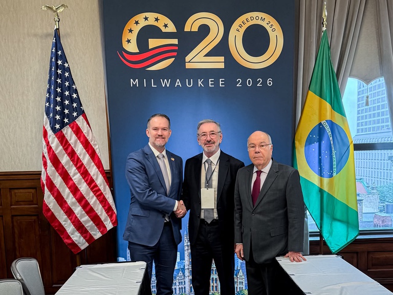 Foto de três autoridades em frente a um painel do evento “G20 Milwaukee 2026”, com as bandeiras dos Estados Unidos e do Brasil ao lado, em ambiente de conferência.