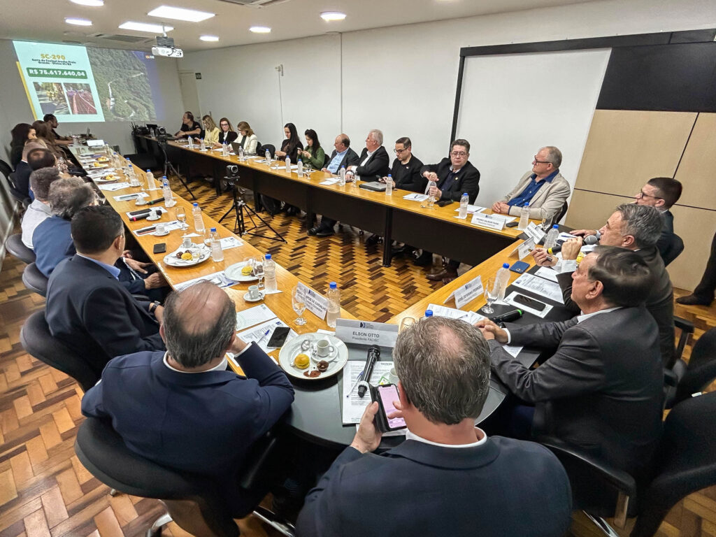 Reunião em sala de conferências com participantes ao redor de uma mesa retangular, documentos e copos sobre o tampo, projetor e telão ao fundo, durante debate