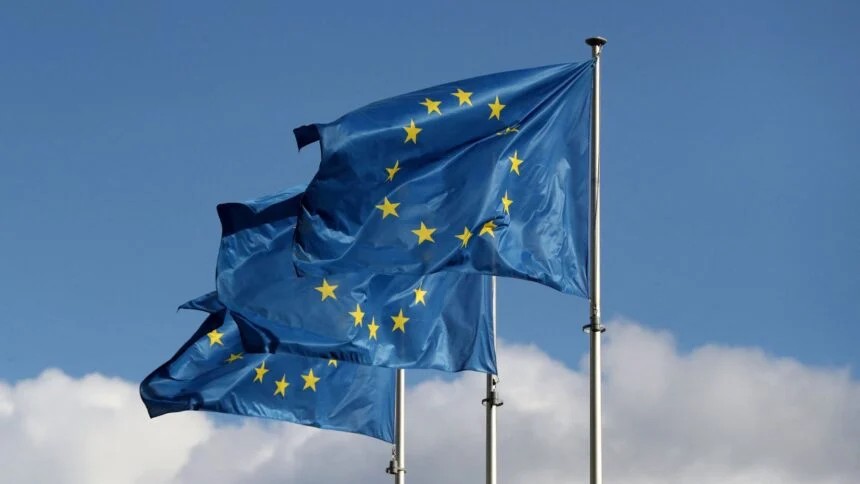Bandeiras da União Europeia azul com estrelas douradas ao vento, hasteadas no céu com nuvens ao fundo.