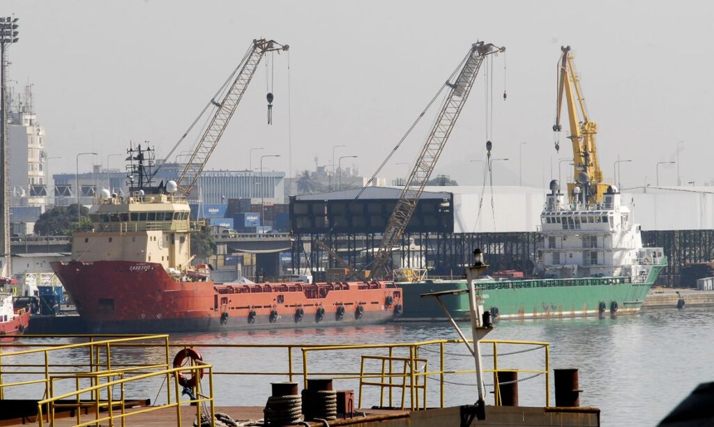 Vista de um porto com navios cargueiros atracados e guindastes de grande porte realizando operações de movimentação de cargas em docas, com céu nublado ao fundo.